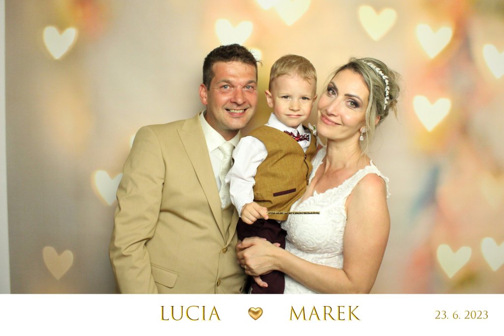 Mladomanželia pózujúci spolu so svojim malým synom pred fotopozadím vo farbach zapadajúceho slnka a žltými srdiečkami.